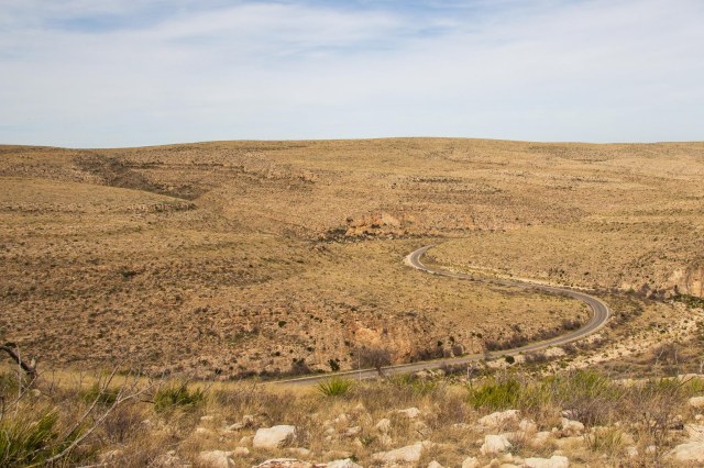 Wispy white clouds against blue sky, rolling golden hills and S-curve road