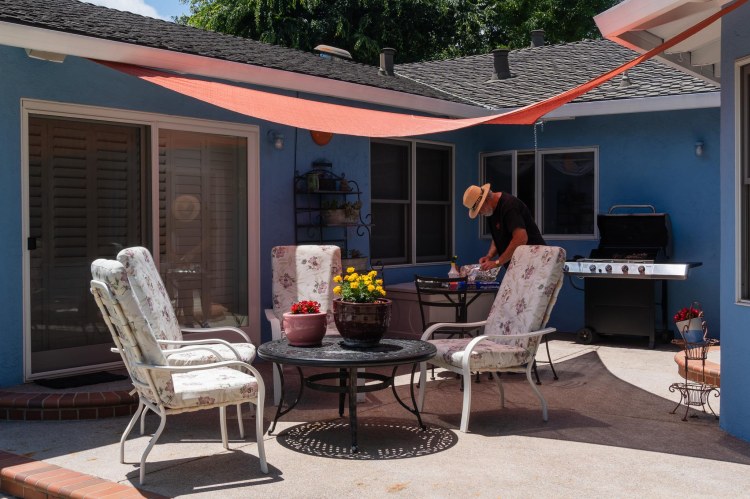 Back of blue house, man cooking, deck chairs, and wrought iron table.