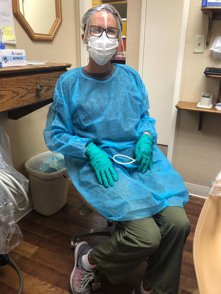 Dental hygienist wearing mask, glasses, protective plastic mask, blue gown, and green gloves