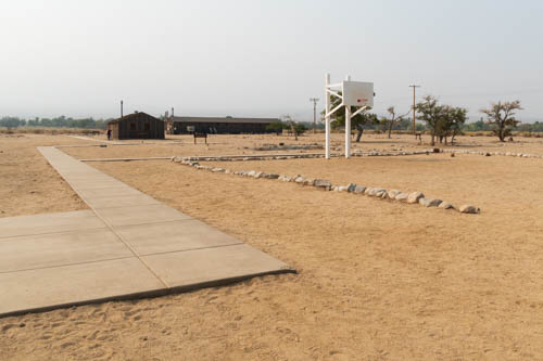 Sidewalk, rocks, sandy area with a few trees and brown buildings in the background.