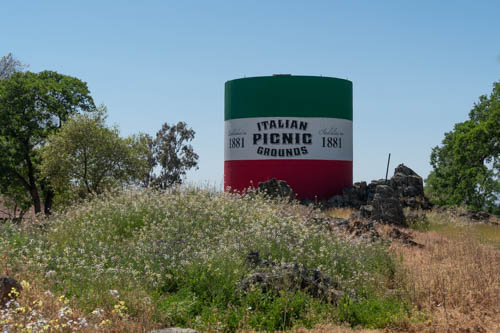 Italian Picnic Grounds green, white and red striped water tower on a hill with green bushes and brown grass.