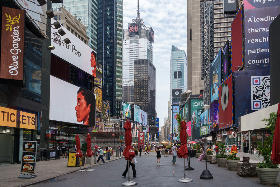 A bustling street view of Times Square in New York City, featuring large digital advertisements, retail stores, and red umbrellas at outdoor seating areas. The sky is overcast, and people are walking along the street.