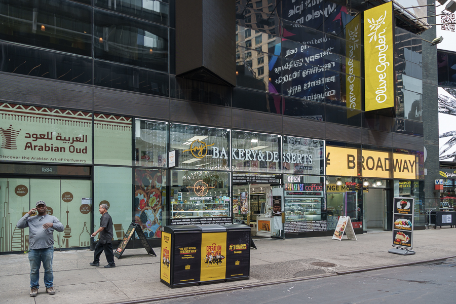 A street view of a city block featuring storefronts for 'Arabian Oud', a bakery and desserts shop, and 'Olive Garden'. Two individuals are walking in front, and various signs advertise services and products.