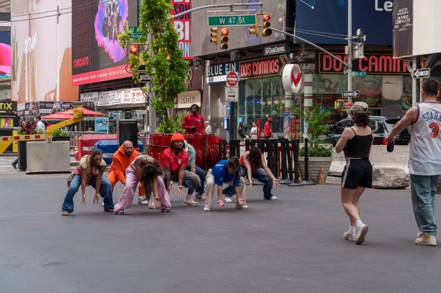 A group of performers in colorful outfits dancing in the street, with Times Square billboards in the background.