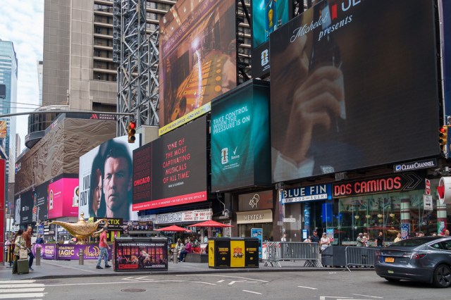 A busy street scene in Times Square featuring large, colorful advertisement billboards and crowds of people.