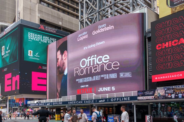 A large digital billboard in Times Square advertising the movie 'Office Romance', featuring actors Jennifer Lopez and Brett Goldstein, with the release date of June 5 and the tagline 'It's Going Down'.