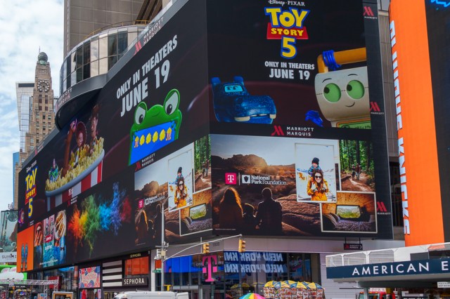 A large digital billboard in Times Square promoting the movie Toy Story 5, featuring colorful graphics and characters from the film, along with details about its release date of June 19.