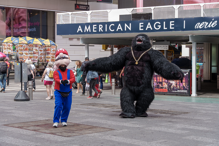 A person dressed as a Mario character stands next to a performer in a gorilla costume, both in a busy urban area with pedestrians in the background.
