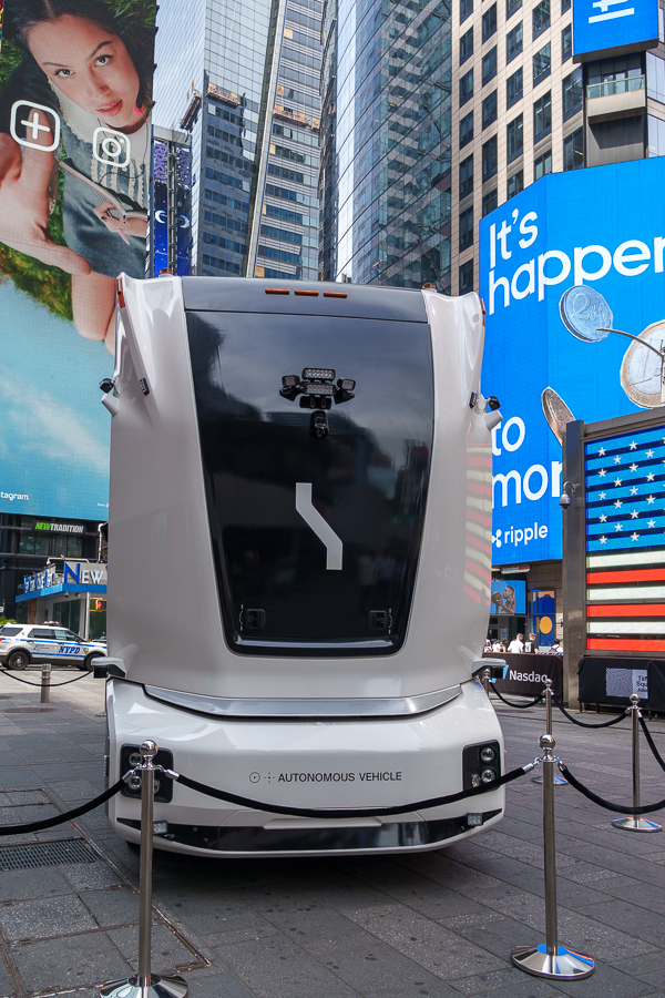 A futuristic autonomous vehicle displayed in Times Square, surrounded by digital billboards and a stanchion barrier.