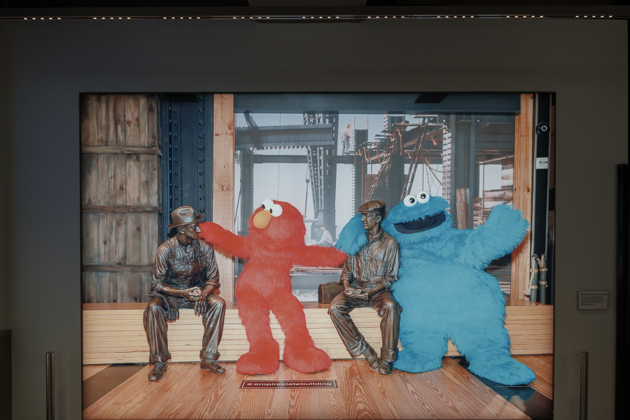 A colorful display featuring Elmo, a red character, and Cookie Monster, a blue character, sitting beside two bronze statues of construction workers in front of a backdrop of a construction site.