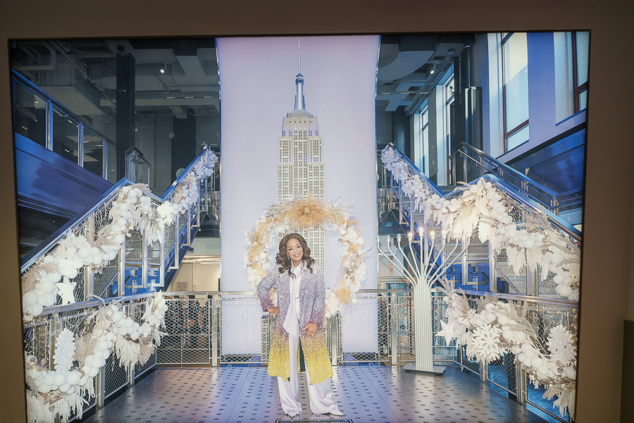 A woman stands confidently in a stylish outfit with a sparkly jacket and yellow pants, framed by decorative white garlands. In the background, the Empire State Building is prominently displayed.
