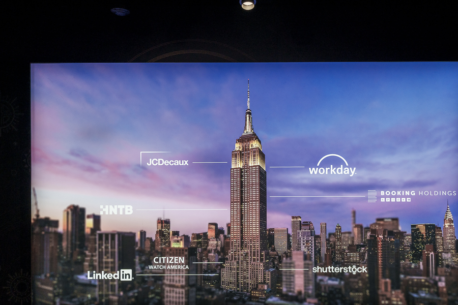 A digital display featuring the Empire State Building with branding logos including JCDDecaux, Workday, Booking Holdings, and others against a sunset skyline backdrop.