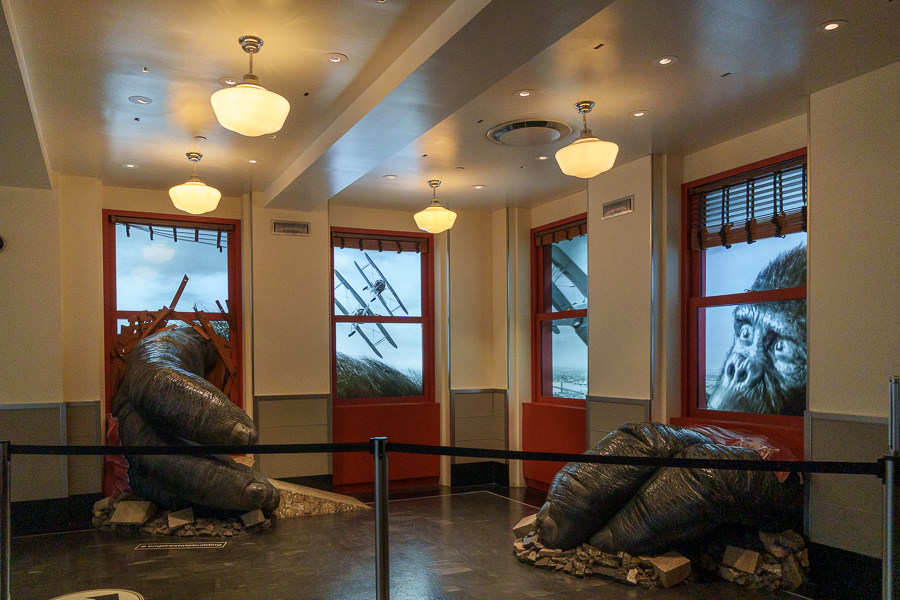 An interior space featuring large gorilla hands emerging from the floor, with a background of windows displaying images of a gorilla's face and planes flying in a stormy sky.
