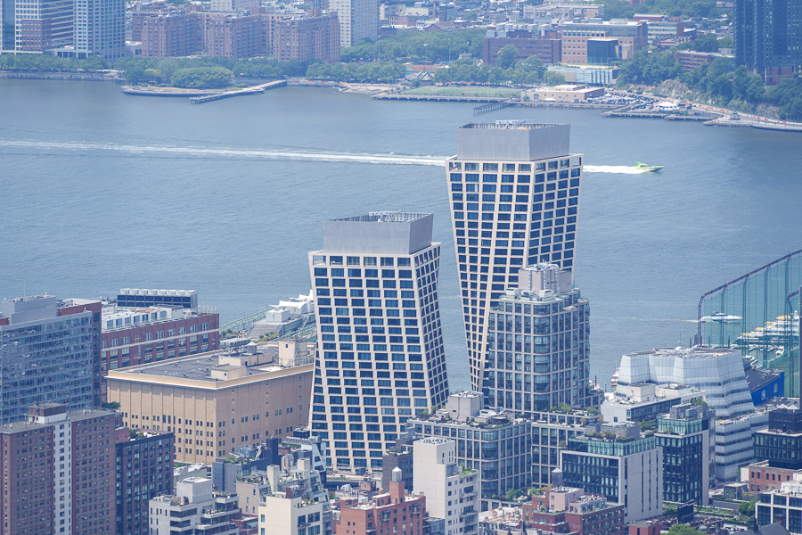 Aerial view of two modern high-rise buildings with a river in the background, surrounded by other urban structures.