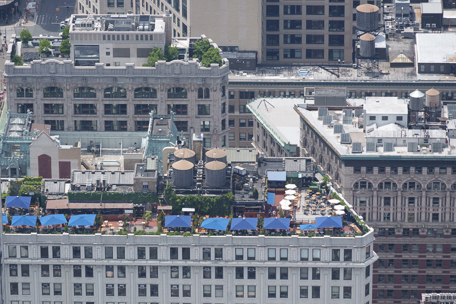 Aerial view of a rooftop terrace featuring blue umbrellas and lounge chairs, surrounded by buildings in an urban setting.