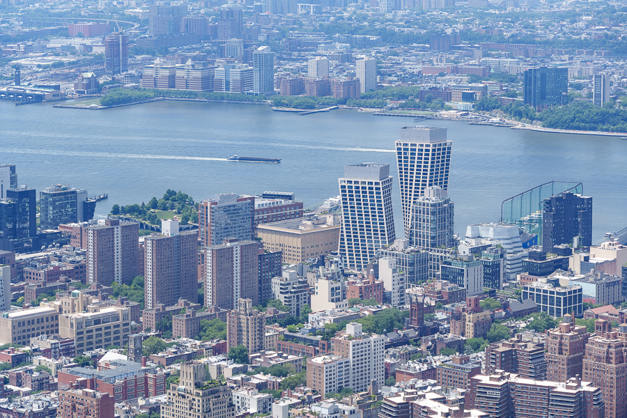 Aerial view of a city skyline featuring various buildings along a river, with a boat traveling on the water and greenery along the waterfront.