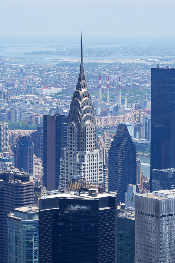 The iconic Chrysler Building towering above the New York City skyline, featuring its distinctive Art Deco spire and intricate architectural details, with a view of the East River and surrounding buildings in the background.