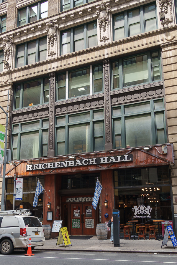 Facade of Reichenbach Hall, featuring large windows and decorative elements, with wooden signage and outdoor seating.