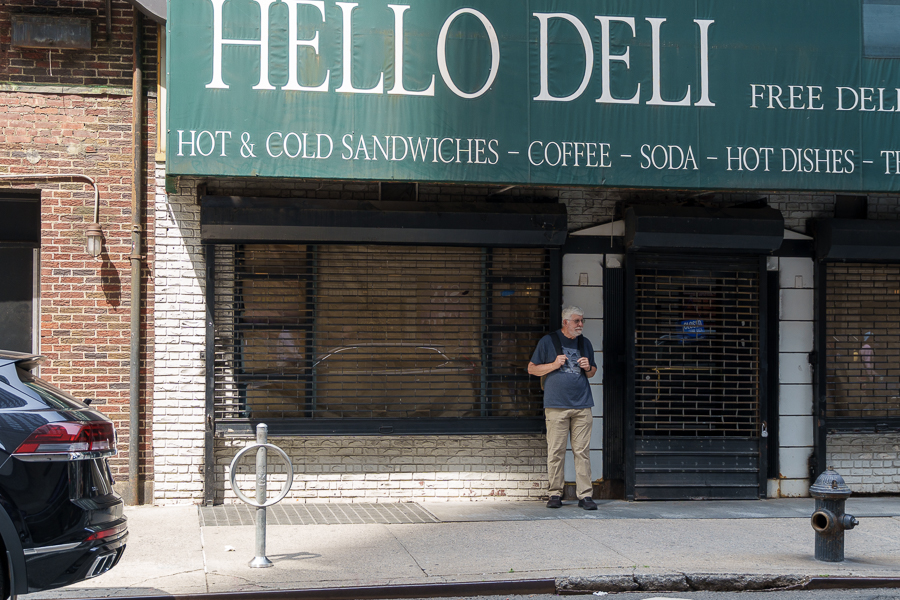 A man stands outside a deli named 'Hello Deli,' which features a large green sign with menu items listed. The shop appears closed with barred windows.