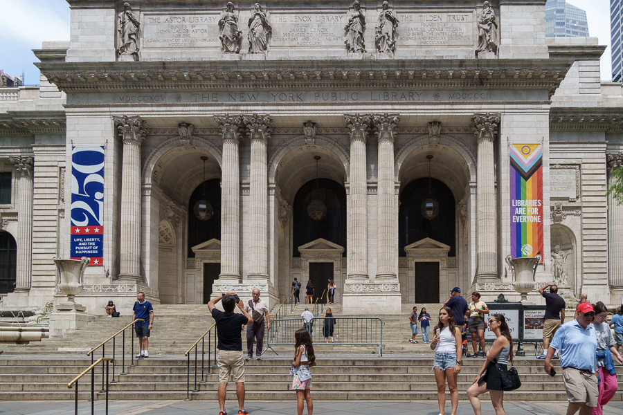 The exterior of the New York Public Library featuring grand columns, banners celebrating its 250th anniversary, and visitors on the steps.