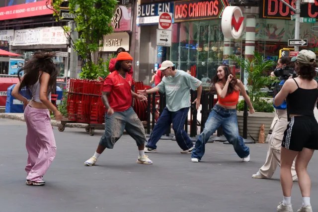 Group of five dancers performing in an urban street setting, with two women in colorful outfits and three men, one wearing an orange hat, surrounded by shops and greenery.